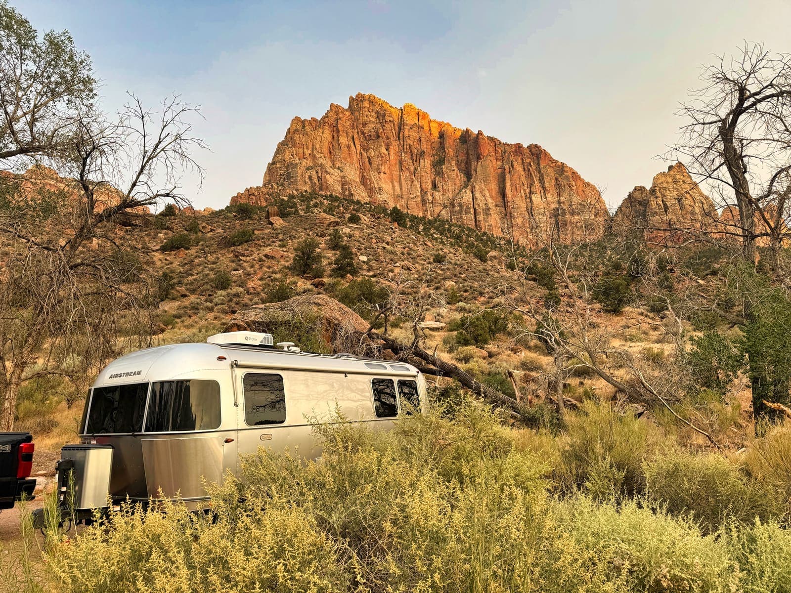 A silver Airstream at camp below a red rock peak in the evening light